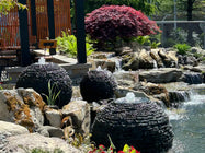 Three dark woven spherical fountain planters with water cascading over rocks in a landscaped garden pond setting.